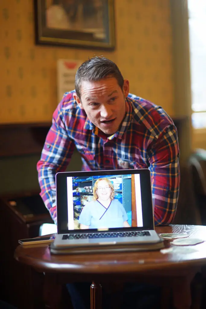 Paul stands behind a laptop computer screen showing a picture of a nurse as he rehearses Paul's Ball