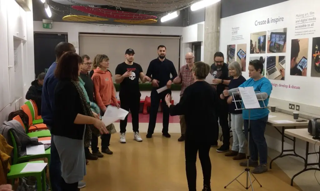 An arts and theatre group standing in a circle holding sheet music, learning a song.